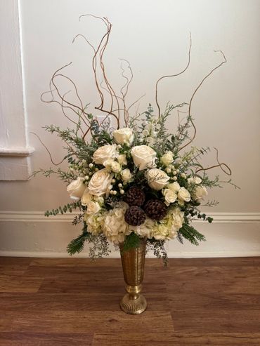 Elegant floral arrangement with white roses and pine cones in a gold vase.