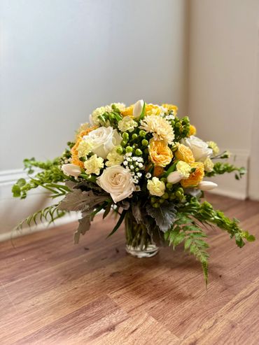 A vibrant bouquet of yellow and white flowers in a glass vase on a wooden floor.