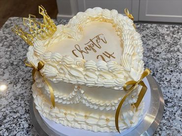 Heart-shaped cake with white frosting, gold ribbons, and a decorative crown on top.