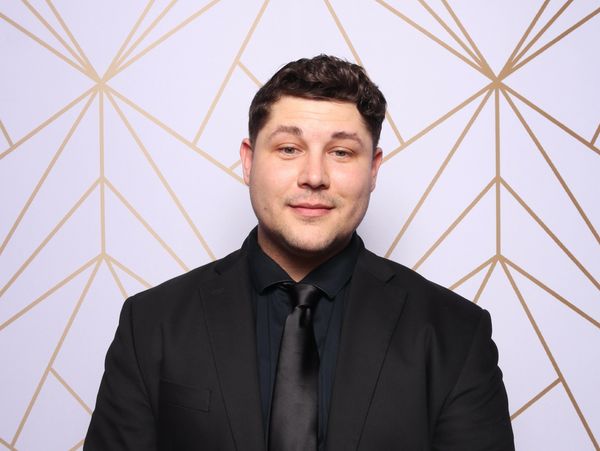 Man in black suit and tie posing against a geometric patterned backdrop.