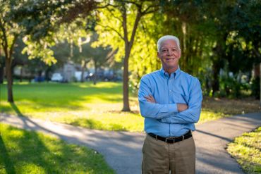 A confident older man standing in a sunny park with arms crossed.