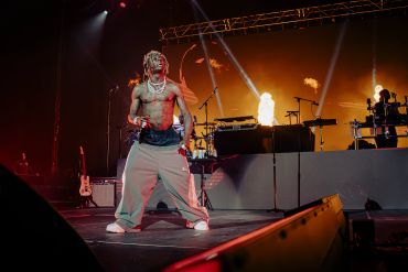 A shirtless performer dances energetically on stage with bright lights and flames behind.
