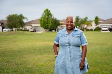 Smiling woman in a denim dress standing in an open grassy area with houses in the background.