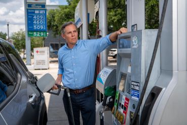 Man in blue shirt refuels car at gas station with high fuel prices shown.