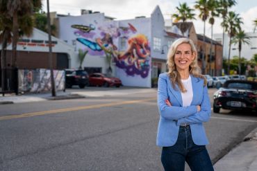Confident woman in blue blazer stands on street with colorful mural behind her.