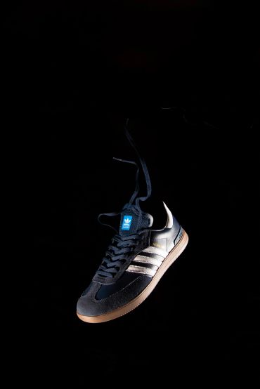 Black Adidas sneaker with white stripes and gum sole against black background.