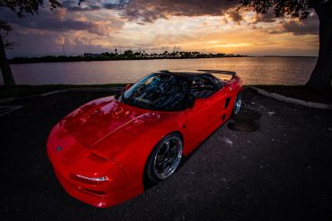 Red sports car parked by a lake during sunset.
