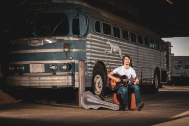 Man playing guitar while sitting on a suitcase near a vintage bus.