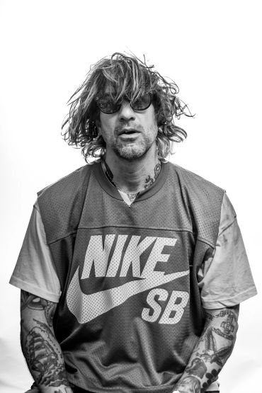 Man with messy hair and tattoos wearing sunglasses and a Nike SB jersey.
