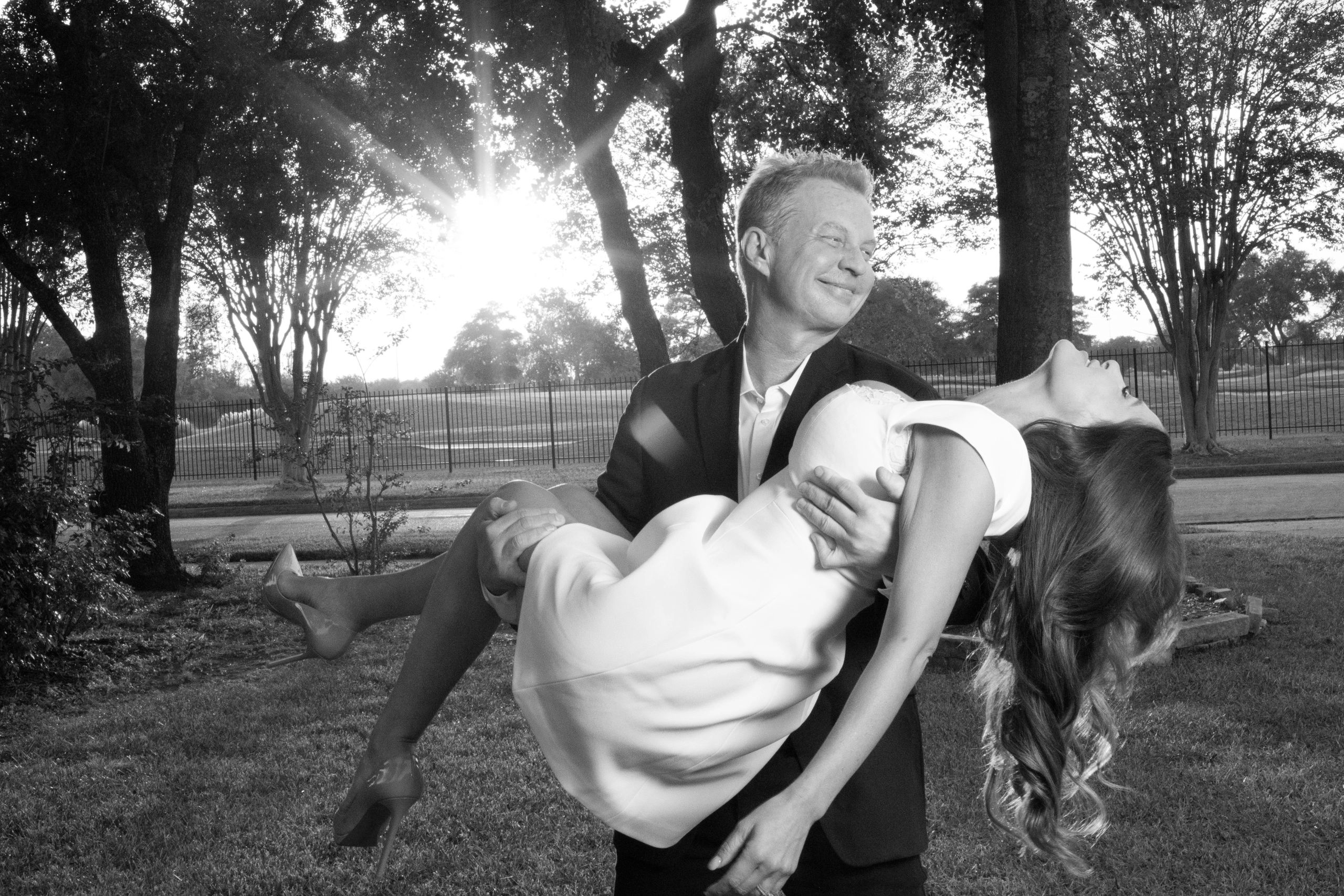 Man in suit holding woman in white dress outdoors at sunset.