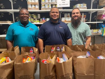 Three men stand behind bags filled with groceries in a food pantry.