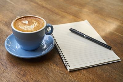 Cup of latte with heart-shaped latte art beside a blank notebook and pen.
