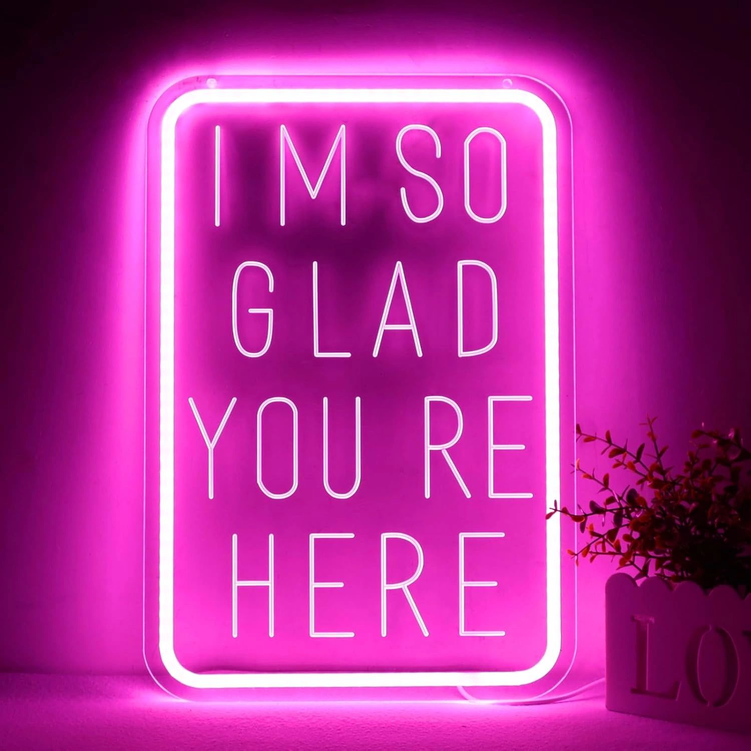 A pink neon sign reads "I'M SO GLAD YOU'RE HERE" glowing brightly.