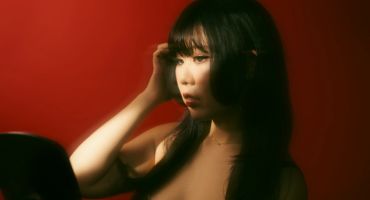 A woman with dark hair looks thoughtfully into a mirror against a red backdrop.