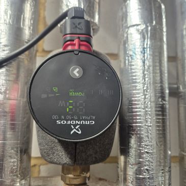 Close-up of a Grundfos Alpha1 circulating pump with digital display on insulated pipes.