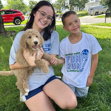 Two kids smiling with a fluffy goldendoodle puppy outdoors.