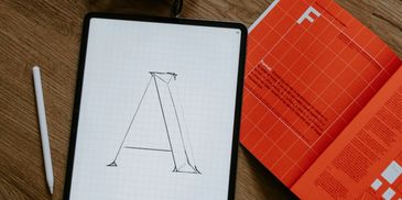 Tablet with letter A sketch, headphones, stylus, and open orange book on wooden floor.