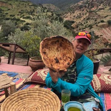 A man proudly holds up a large round flatbread outdoors in a mountainous setting.