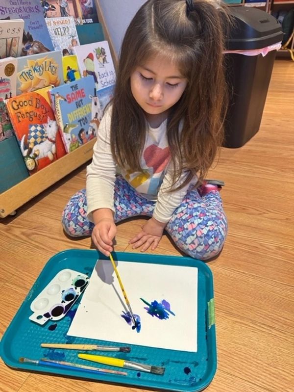 a child sitting on the floor using blue water colors on white piece of paper