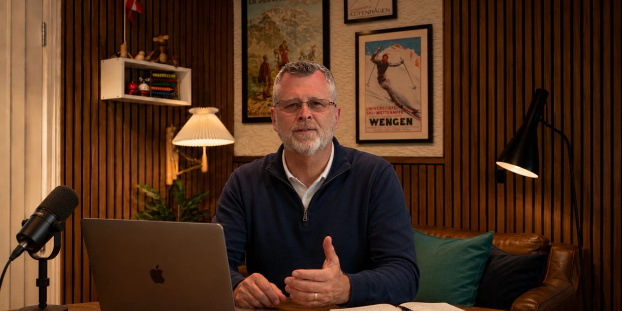 Man speaking in a cozy, wood-paneled room with laptop and microphone.