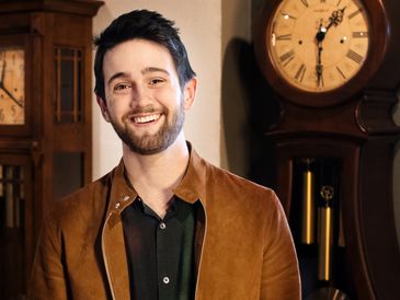 A young man standing in front of a grandfather clock in a suede jacket.