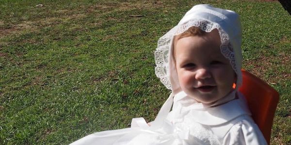 Baby in white bonnet and dress sitting outdoors on grass.