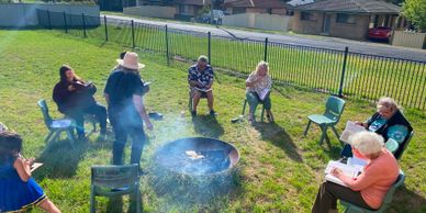 People sitting in a circle outdoors around a fire pit on a sunny day.