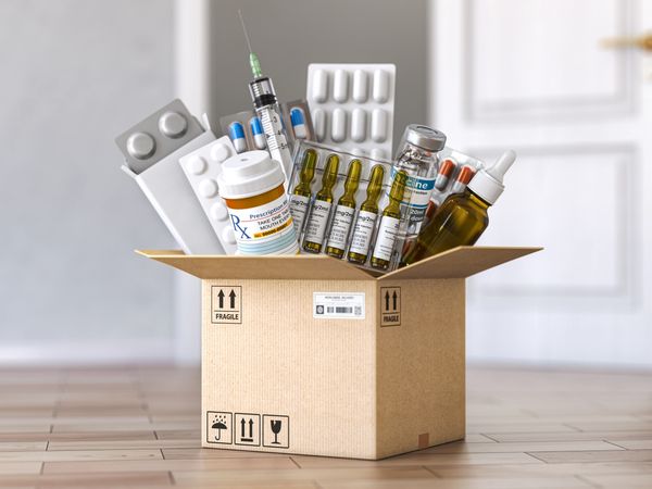 A cardboard box overflows with various medical supplies, including vials, pills, and a syringe.