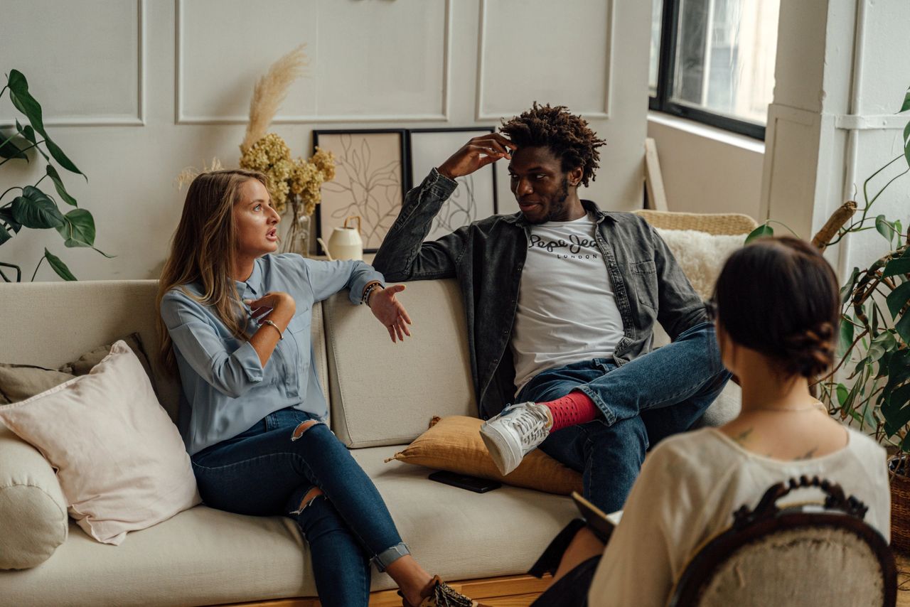 A woman and man sitting on a couch looking at each other while a counselor sits across from them.
