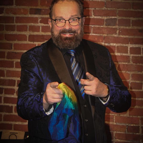 Man with glasses and beard points while holding a colorful cloth against a brick wall.