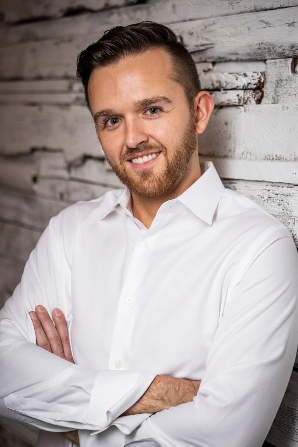 Smiling man in white shirt leaning against rustic wooden wall.