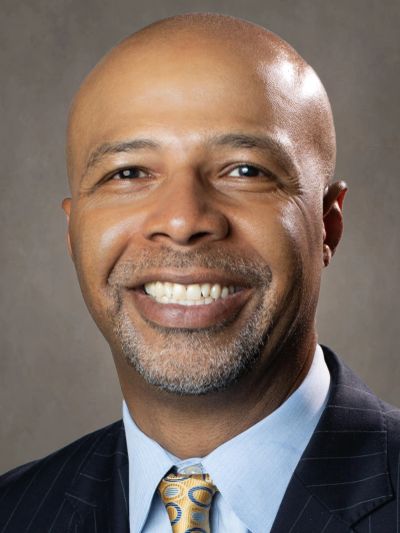 Smiling man in suit with patterned tie against a neutral background.
