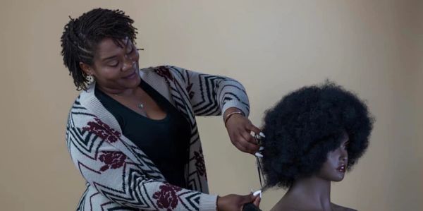 Photo of  the owner Teme Battle, cutting hair on a mannequin, smiling while wearing a sweater.