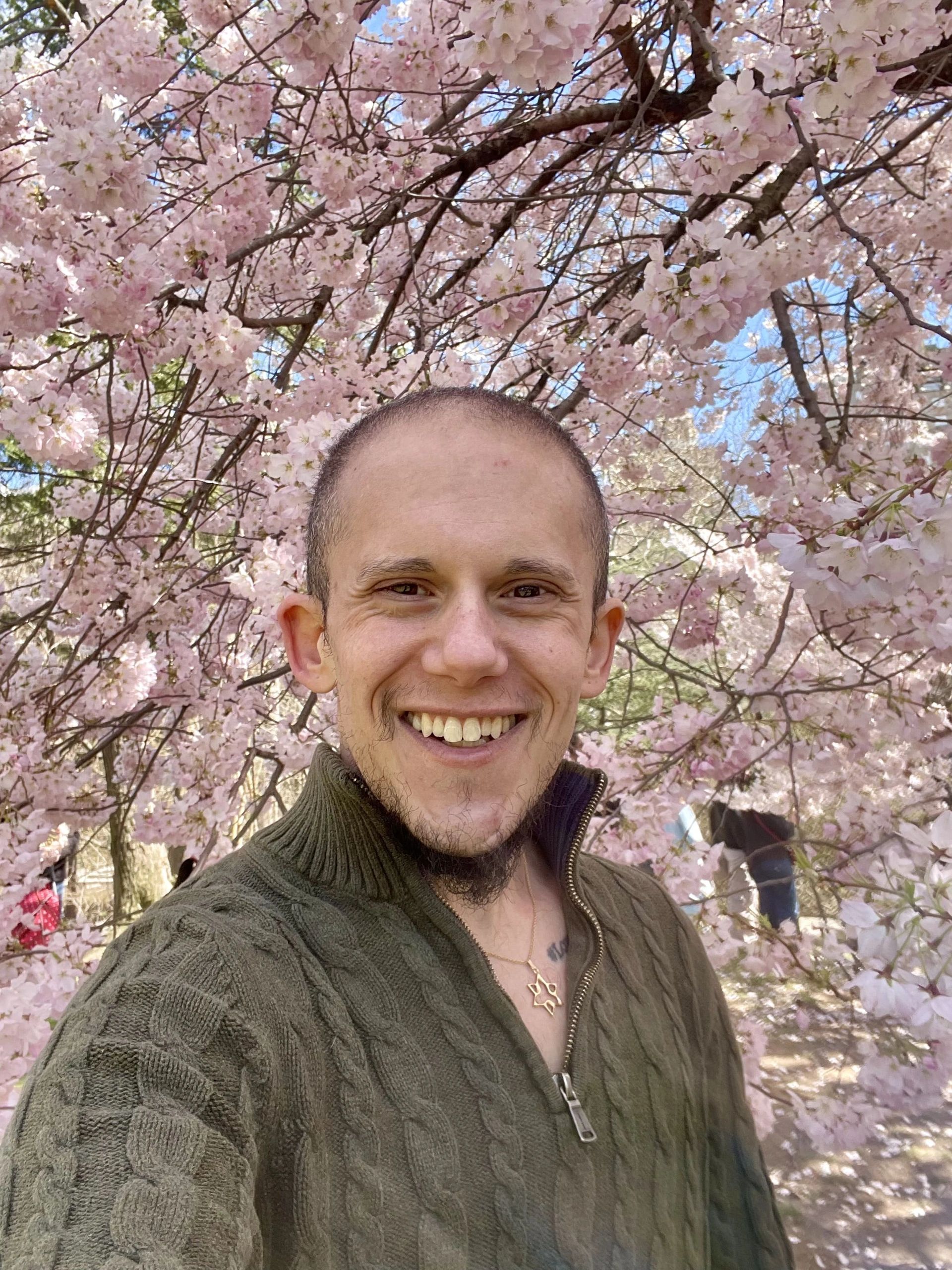 Jamie smiling in dark olive green sweater slightly unzipped, chest-up, in front of cherry blossoms