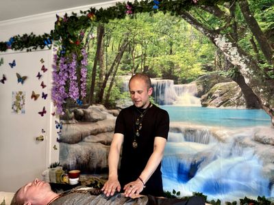 Jamie providing Reiki to client on table in black shirt, in front or rainforest waterfall tapestry