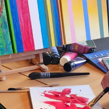 Painting supplies and colorful canvases on a table with someone painting.