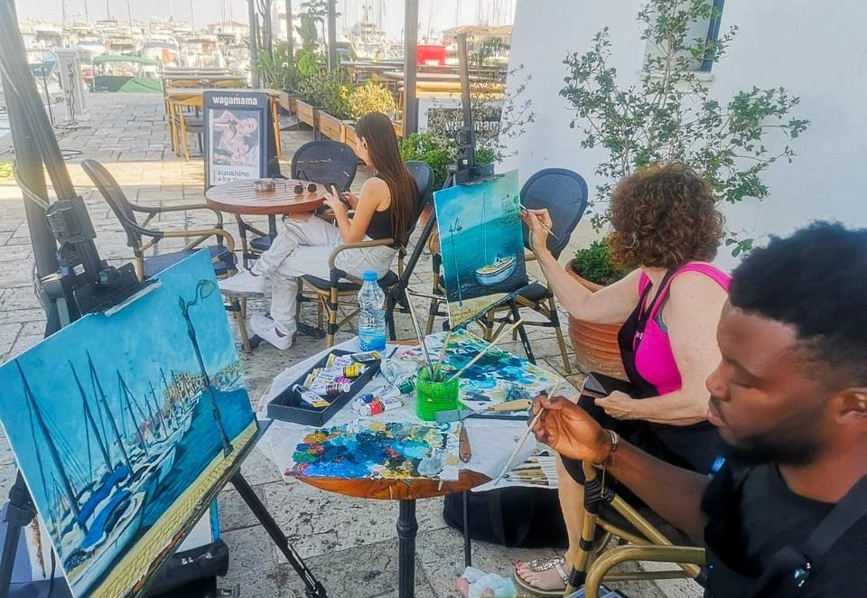 Three people painting maritime scenes outdoors near a harbor.