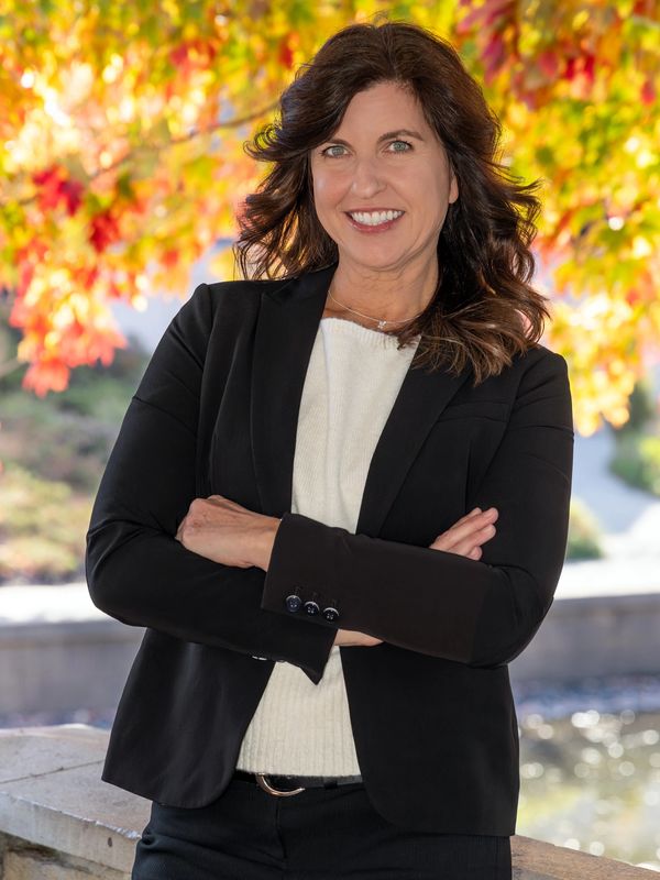 Confident woman in black blazer smiling outdoors with autumn leaves in background.