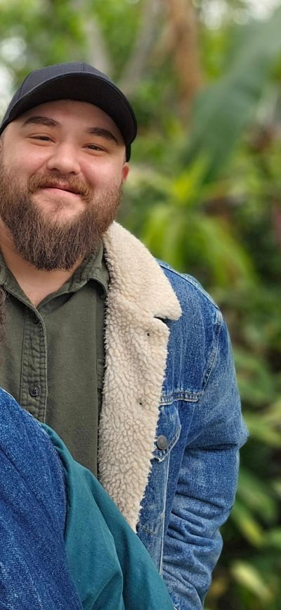 Man wearing a denim jacket and cap smiling outdoors.