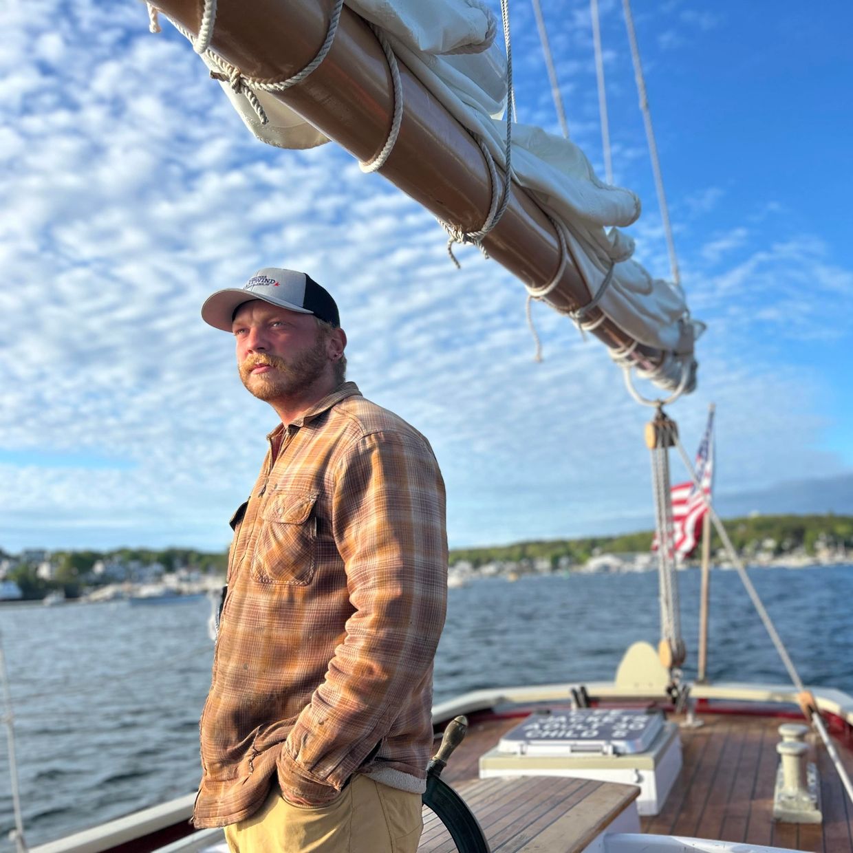 A man in a cap and plaid shirt stands on a boat under a blue sky.