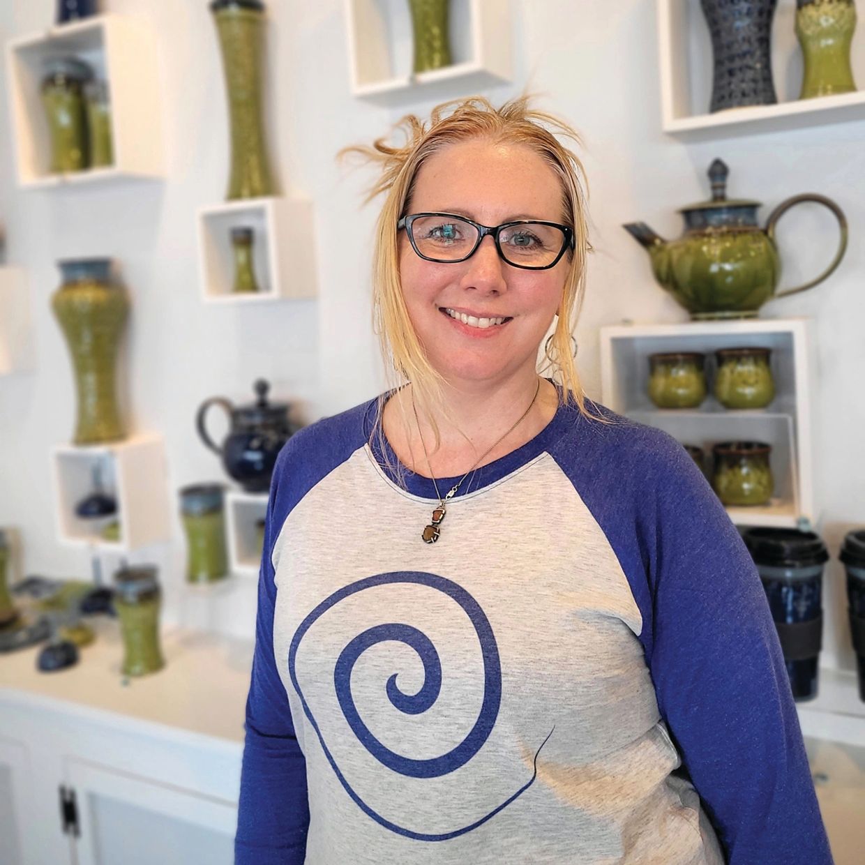 Kelsey Schissel standing in her gallery.  Her handmade pottery is in the background. 