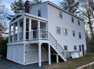 White two-story house with a raised porch and stairs in a wooded area.