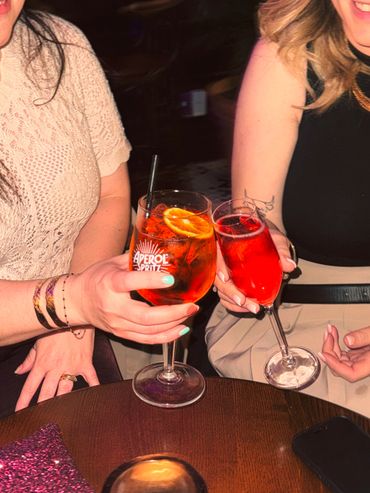 Two women toasting with colorful cocktails at a dimly lit bar.