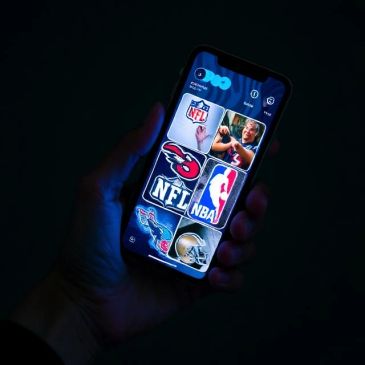 Hand holding a smartphone displaying sports logos in a dark setting.