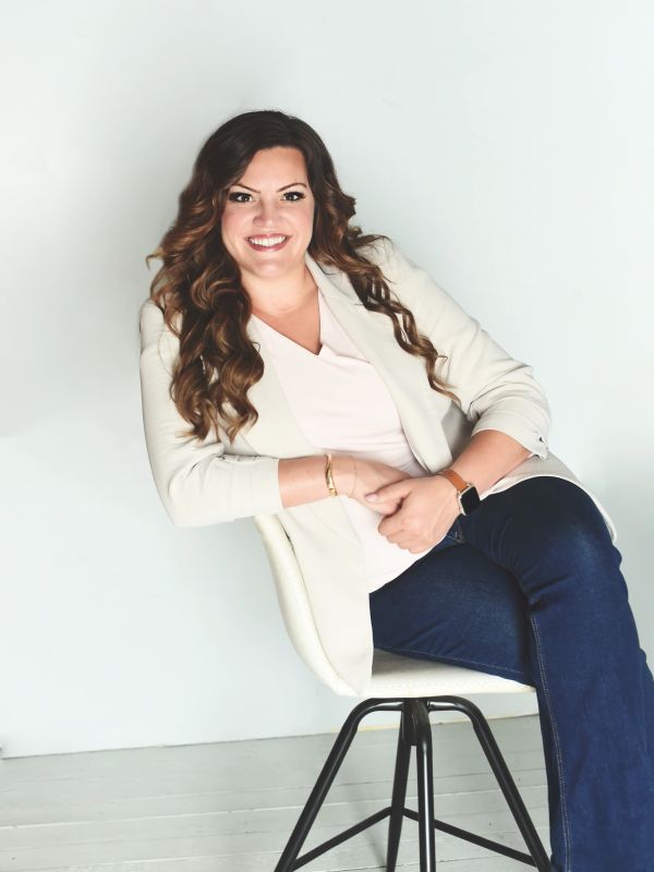 Smiling woman in casual professional attire sitting on a chair against a plain background.