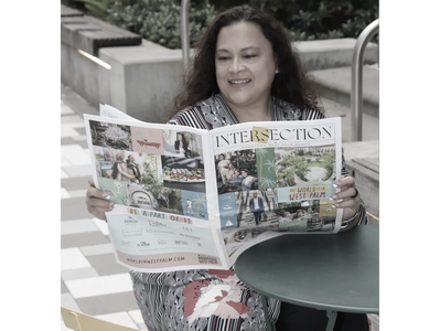 Woman smiling while reading a newspaper outdoors at a table.