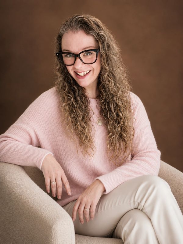 Smiling woman with curly hair, glasses, and a pink sweater sitting on a beige chair.