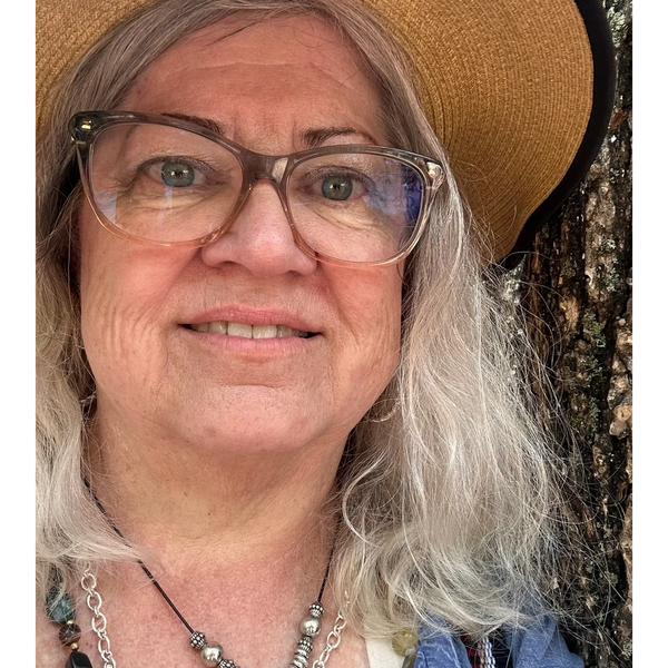 Portrait of a smiling woman with silver-gray hair, glasses, and a tan sun hat standing beside a tree