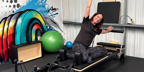 Woman exercising on a Pilates reformer machine, smiling and stretching one arm up.