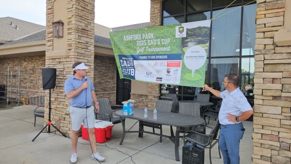Two men at Ashford Park 2025 Dad's Cup Golf Tournament with banner and microphone.